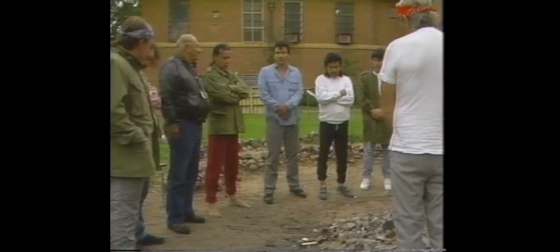 a group of men along with James "Jimmy" Jackson stand in a circle outdoors in front of a prison building. 