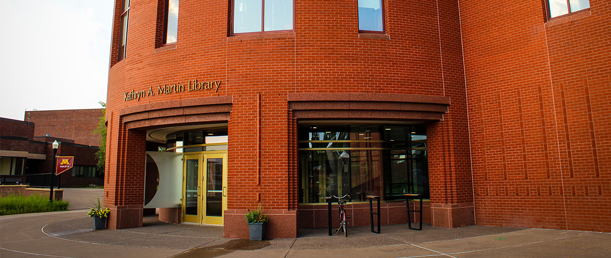 Entrance of Kathryn A. Martin Library, featuring a round brick façade, golden double doors, and a bicycle at the entrance, set within a campus environment.