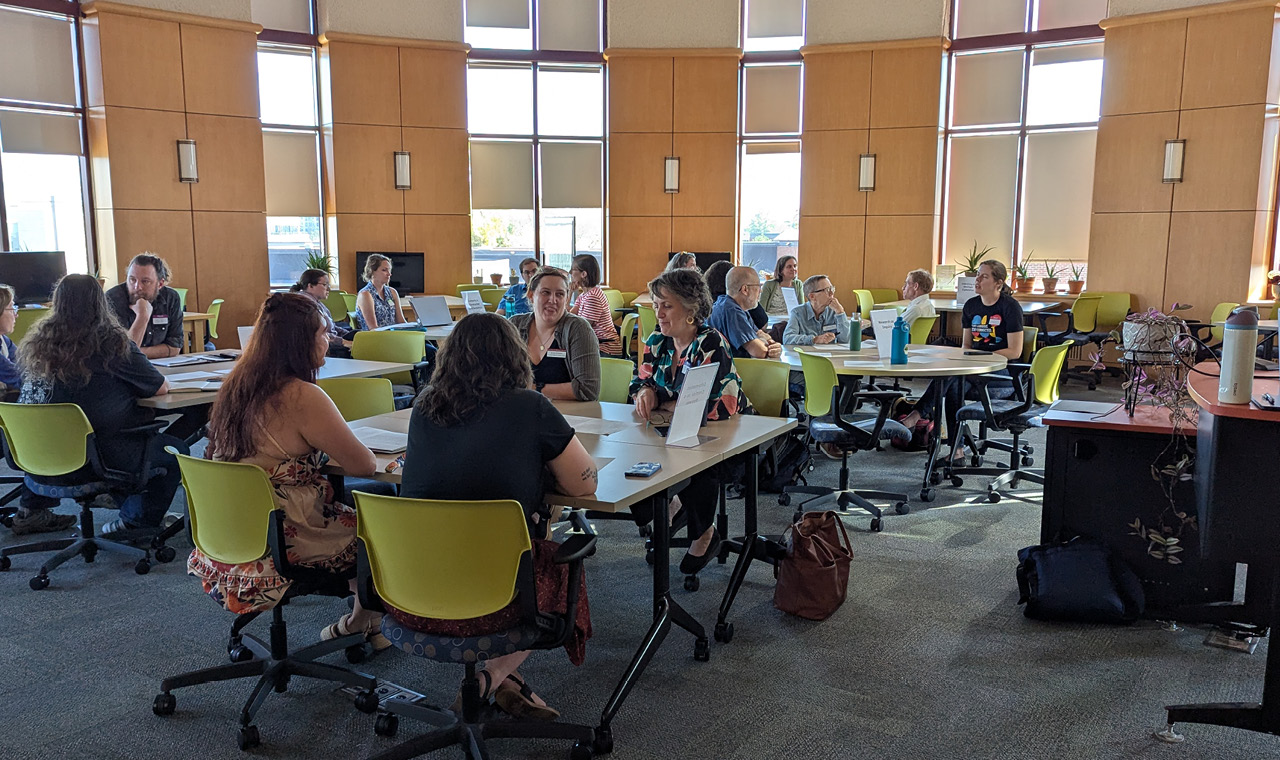 Adults participate in a group discussion around tables in a bright, windowed meeting room.