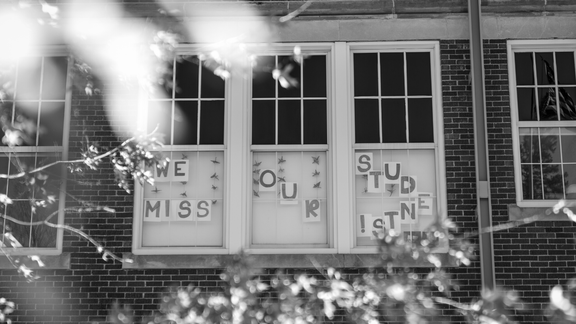 a black and white image of an exterior window at a school. "we miss our students" is posted in the windows.