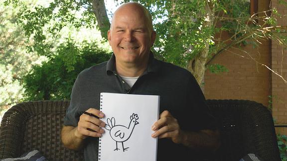 artist Brian Barber sits outside on a summer day, he holds a sketch book with a drawing of a cartoon chicken on it