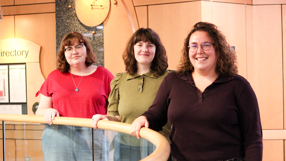 a photograph of three women standing by a glass railing. the photograph is taken on the second floor of the library.. 