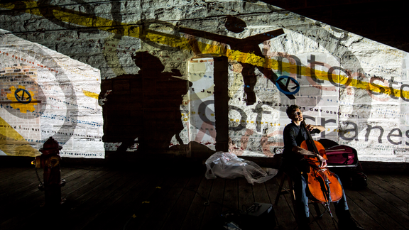 A high contrast photograph of artist Kathy McTavish with her cello, behind her is a wall featuring words and shapes.