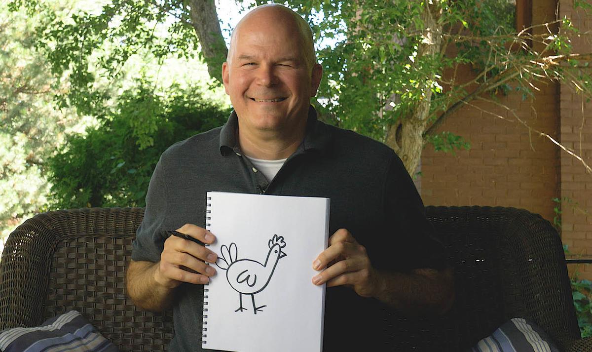 artist Brian Barber sits outside on a summer day, he holds a sketch book with a drawing of a cartoon chicken on it