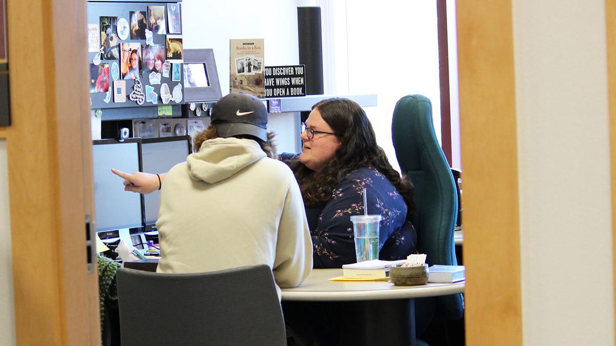 a student sits in an office with a librarian. the student is receiving research help.