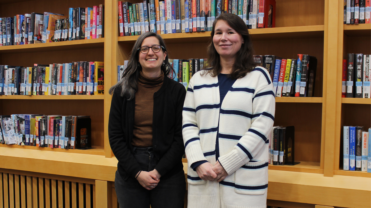 a photograph of two library staff (Kayleen Jones and Cassandra Prang) in front of the book shelves on first floor of the library